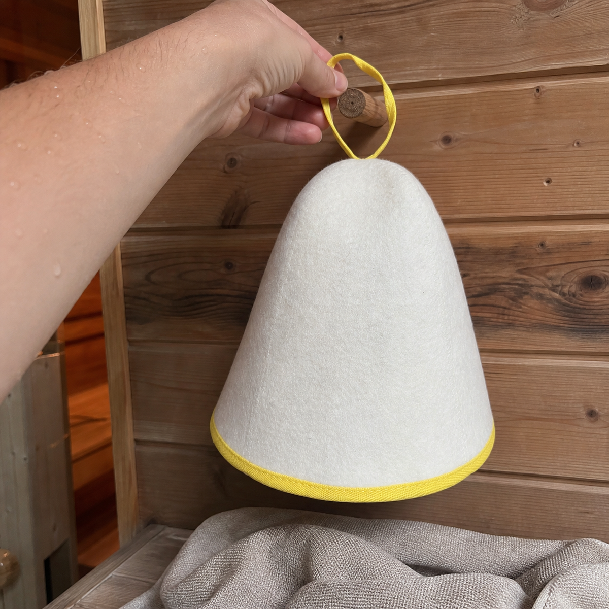 Brand-created UGC-style photo of the white felt sauna hat with yellow trim being hung by its loop